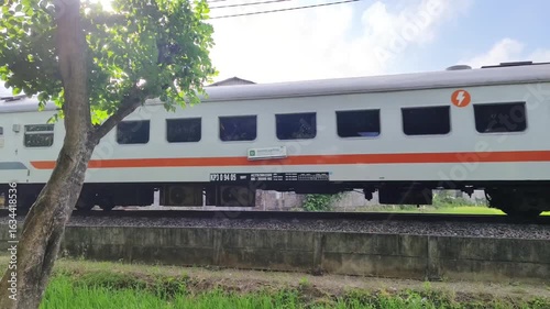 A white and red KAI Ekonomi train is seen passing by on a track, with lush green rice fields in the foreground and buildings; Kediri, Indonesia- August 07, 2025