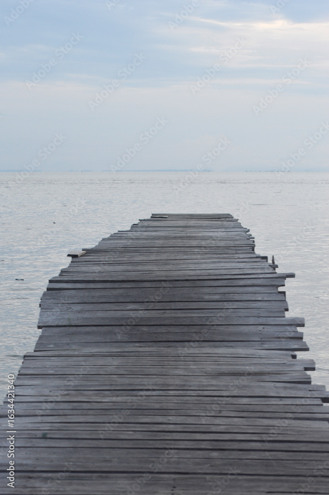 Obraz premium Serene weathered wooden pier leading to calm ocean waters under a vast, pale sky