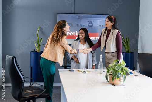 Diverse female colleagues in suits shaking hands across table at office meeting with display
