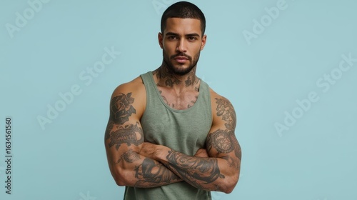 Muscular tattooed man with arms crossed posing against a light blue background