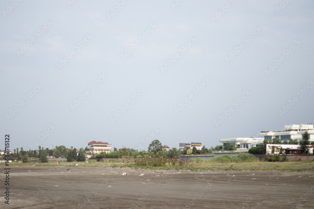 Fototapeta premium Distant view of a coastal neighborhood with modern villas arranged along a low shore. A large pale sky occupies much of the frame, placing the houses as a thin band above the empty foreground