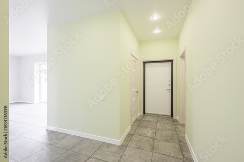 modern hallway with pale green walls, tiled floors, and white doors under recessed lighting. An open doorway leads to a bright room
