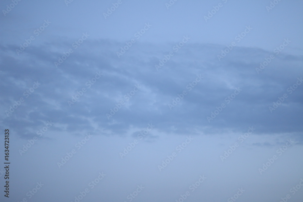 Obraz premium Calm evening sky with a long smooth cloud bank drifting across a pale blue background, photographed without ground elements to emphasize the atmospheric form. Soft illumination creates delicate
