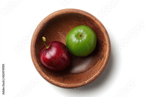 A dark brown wooden bowl holds a vibrant red cherry and a green, unripe counterpart against a stark white backdrop, viewed from directly above