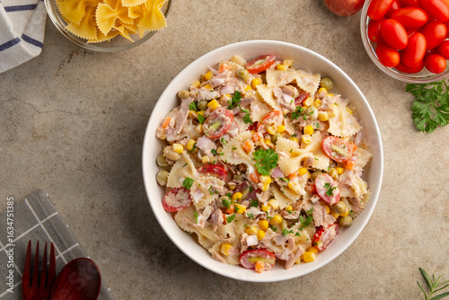 Homemade macaroni salad with bow tie pasta and canned tuna, carrot, onion, corn, tomato, green peas and mayonnaise dressing in a white plate on wooden table.Top view.