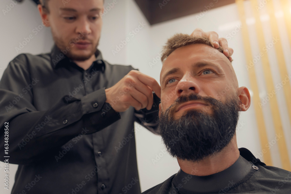 Fototapeta premium A skilled hairdresser is shaping a man's bald fade haircut and trimming his beard in a contemporary barbershop. The atmosphere is relaxed, focused on grooming and style