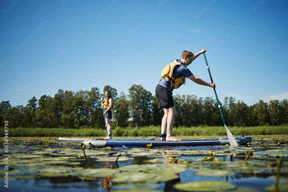 Naklejka premium Caucasian young adult man and Caucasian young adult woman paddleboarding on lake covered with water lilies, both wearing life vests, standing on separate boards, trees in background