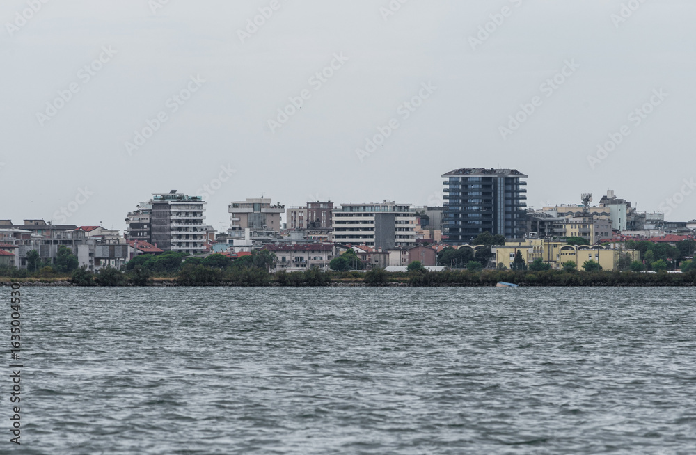 Obraz premium Grado, Italy - July 24, 2025: Panoramic view of Grado from Barbana island, with modern buildings rising above the lagoon on a cloudy day over calm northern waters.