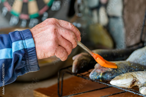 Foto Close-up of a senior man's weathered hand using an orange silicone brush to baste fresh trout on a barbecue grill