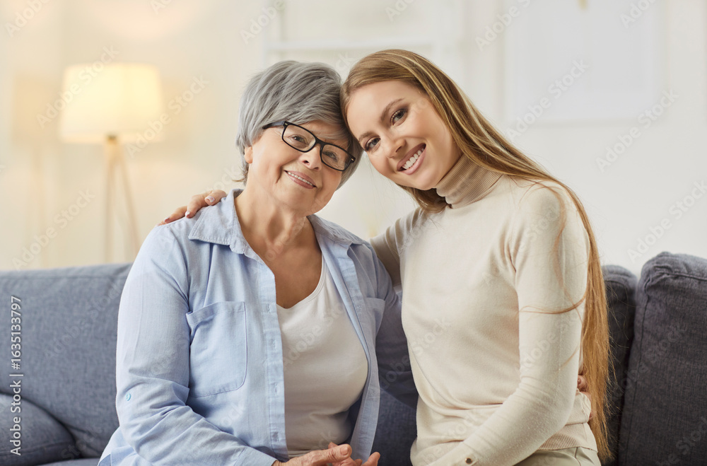 Smiling elderly and young women sitting on cozy sofa, embracing and looking at camera. Happy senior mother and her adult daughter hugging, showing love, bond and warmth shared between two generations. Smiling elderly and young women sitting on cozy sofa, embracing and looking at camera. Happy senior mother and her adult daughter hugging, showing love, bond and warmth shared between two generations.