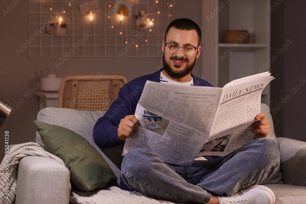 Fototapeta premium Young man with eyeglasses reading newspaper on sofa at home in evening