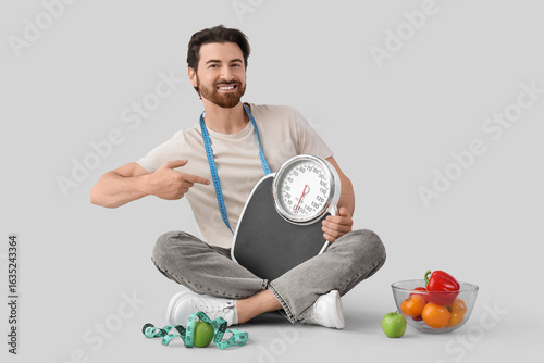 Young man pointing at weight scales on light background