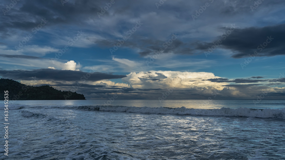 Obraz premium Early morning. Twilight. The ocean waves roll towards the shore, foam, and spread out. The clouds in the blue sky are illuminated with golden light. A hill in the distance. Malaysia. Borneo. 