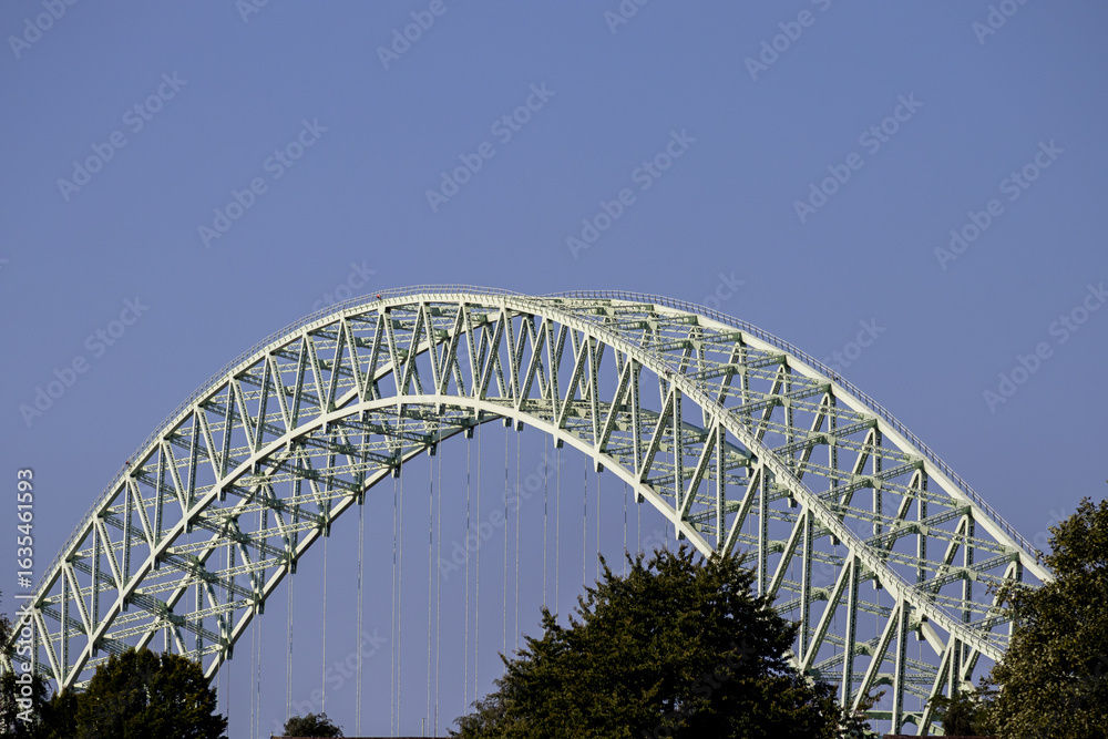 Fototapeta premium Steel arch of the Silver Jubilee Bridge rises above treetops, framed by a cloudless blue sky in bright sunlight
