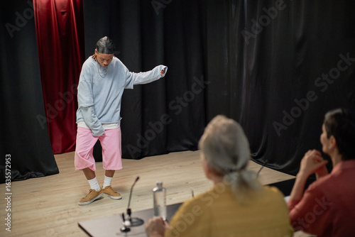 Man performing dance routine in front of two judges during talent competition, judges observing and evaluating performance, stage with black curtains in background