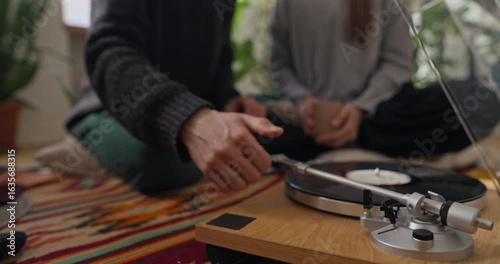Close up of a guy putting a vinyl record in a player and turning it on while listening to vintage music with his girlfriend and having tea at home in a cozy apartment