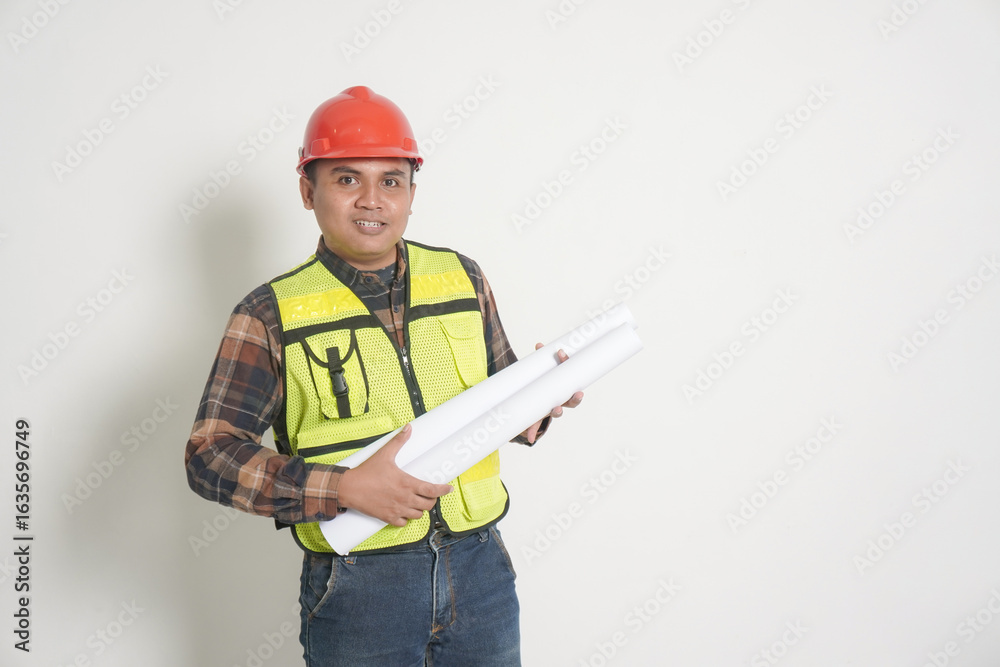 Fototapeta premium Asian construction worker wearing safety vest and helmet, holding project paper plans. Isolated image on white background