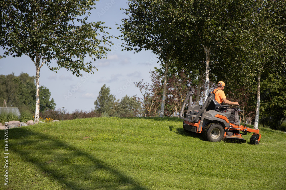 Fototapeta premium Man Operating Lawn Mower in Sunny Park Landscape with Green Grass