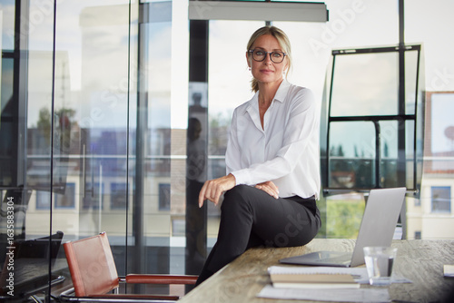 Confident professional sitting on desk in modern office