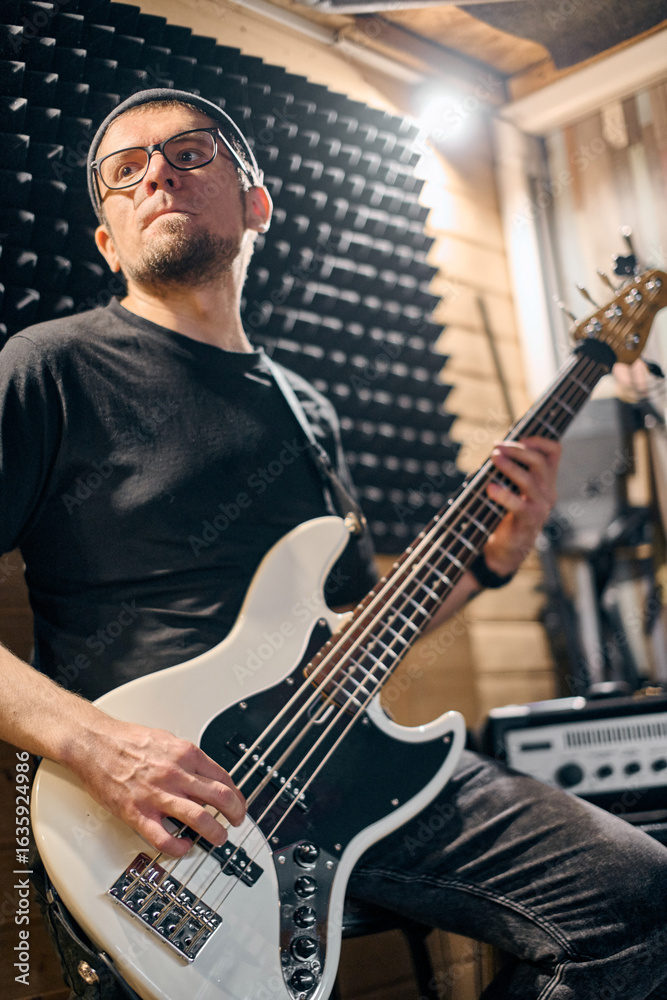 Fototapeta premium Young adult male with glasses playing bass guitar passionately in soundproof studio. Dim lighting and wooden walls create intimate rehearsal environment. Focused expression suggests dedicated practice