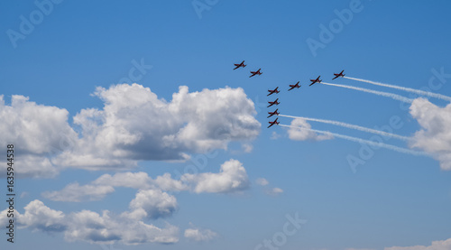squadron of nine jet planes performs an aerial formation against a bright blue sky, leaving white trails behind