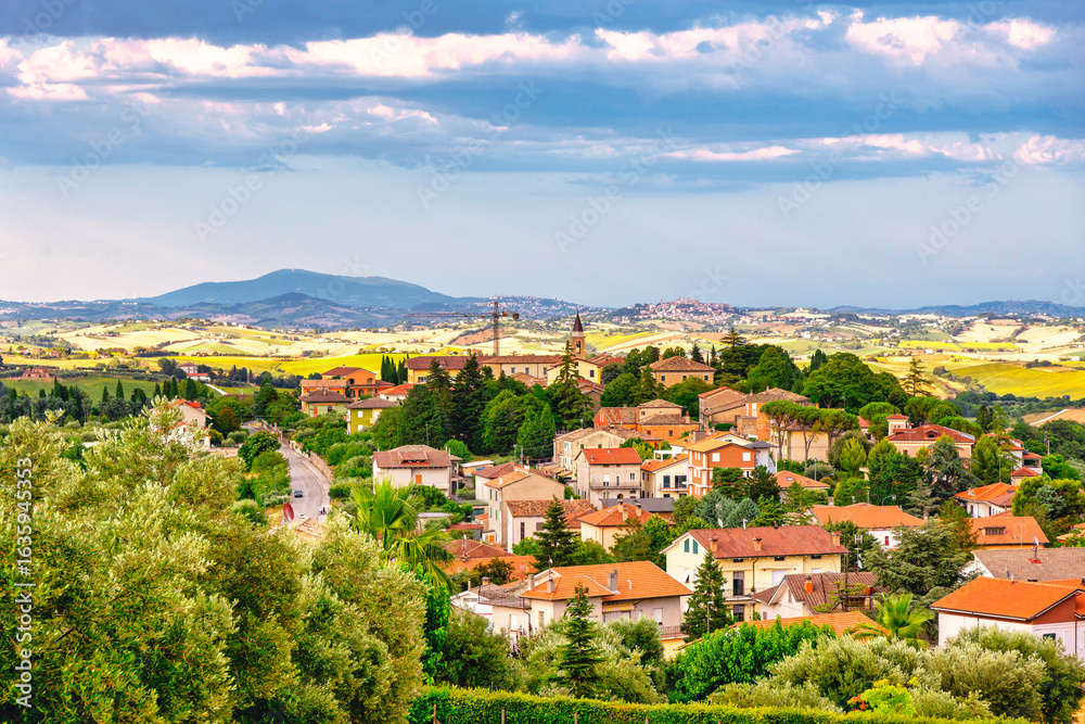 Fototapeta premium Charming hilltop village with red-roofed houses, winding road, and church steeple, surrounded by lush greenery and distant mountains under a partly cloudy sky.