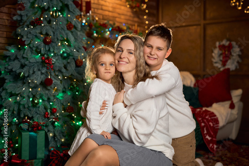 smiling mother and her two children in white sweaters hug closely, celebrating in a cozy, festive room with a decorated tree and golden lights