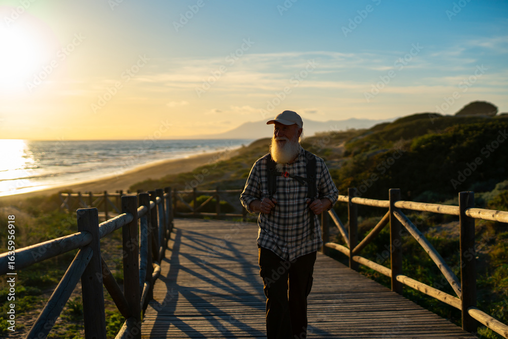 Fototapeta premium Senior Man with a White Beard Walking Along a Scenic Coastal Boardwalk at Sunset, Wearing a Plaid Shirt and a Backpack, Enjoying the Tranquility of Nature