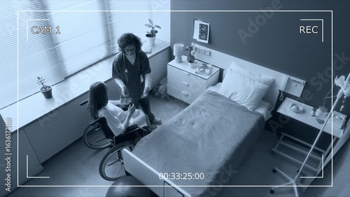 Wide top view of CCTV security camera filming female nurse transporting heavily pregnant woman in wheelchair to maternity ward in hospital, helping into bed while talking