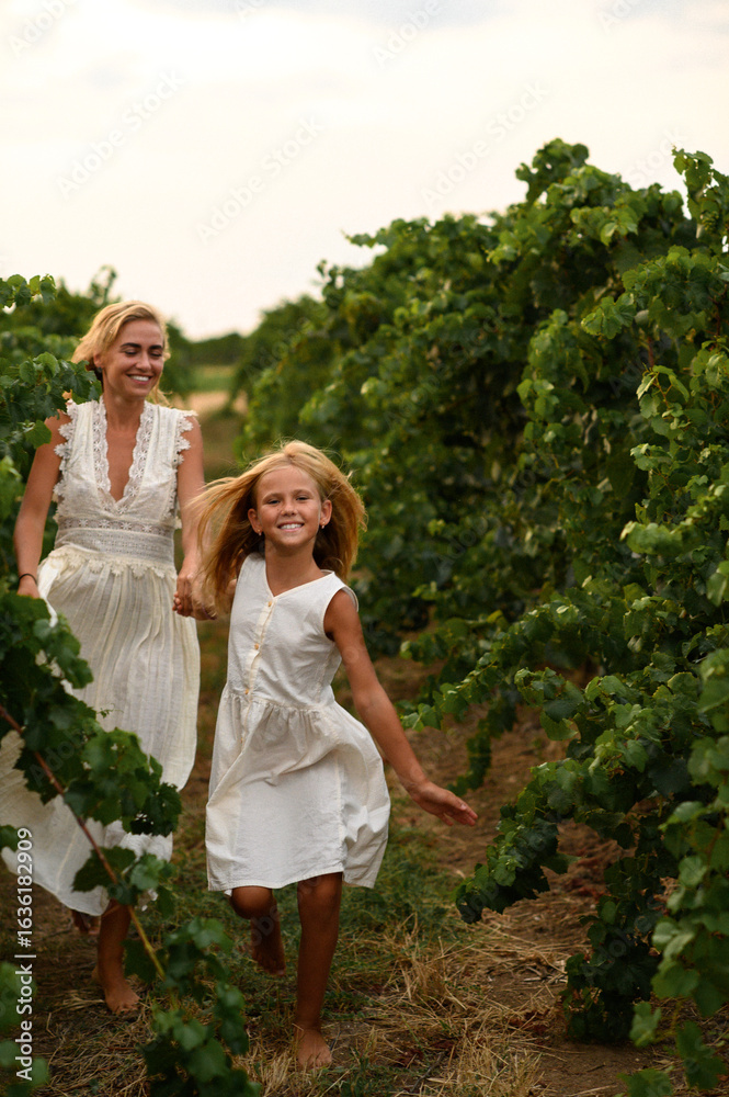 Fototapeta premium Mother and daughter in white dresses running and laughing through a sunny vineyard, enjoying family time, freedom and happiness in nature.