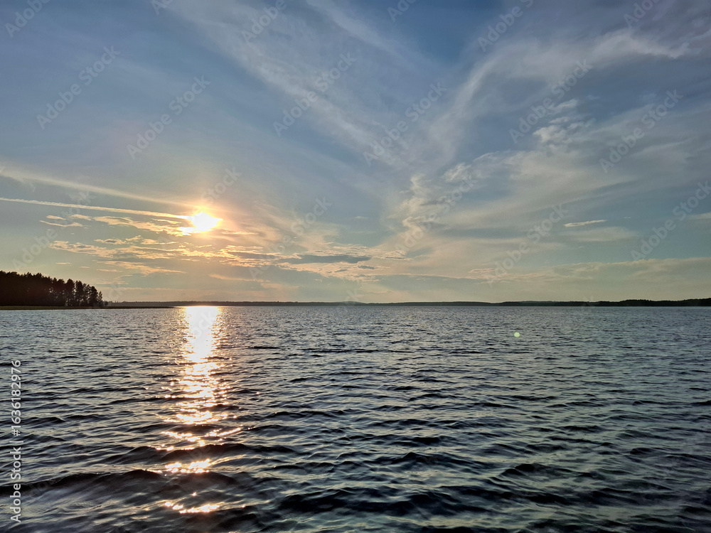 Fototapeta premium Golden sunset over a peaceful lake with rippling water and a glowing sky. The silhouette of a forest lines the horizon, creating a serene and scenic summer evening atmosphere.