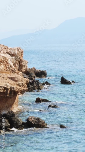 Wallpaper Mural Scenic rocky seashore of Aegina island, Greece. Tall light cliff with dark boulders at the base, water near the shore turning deep blue to horizon, distant mountains in hazy summer light, no people Torontodigital.ca