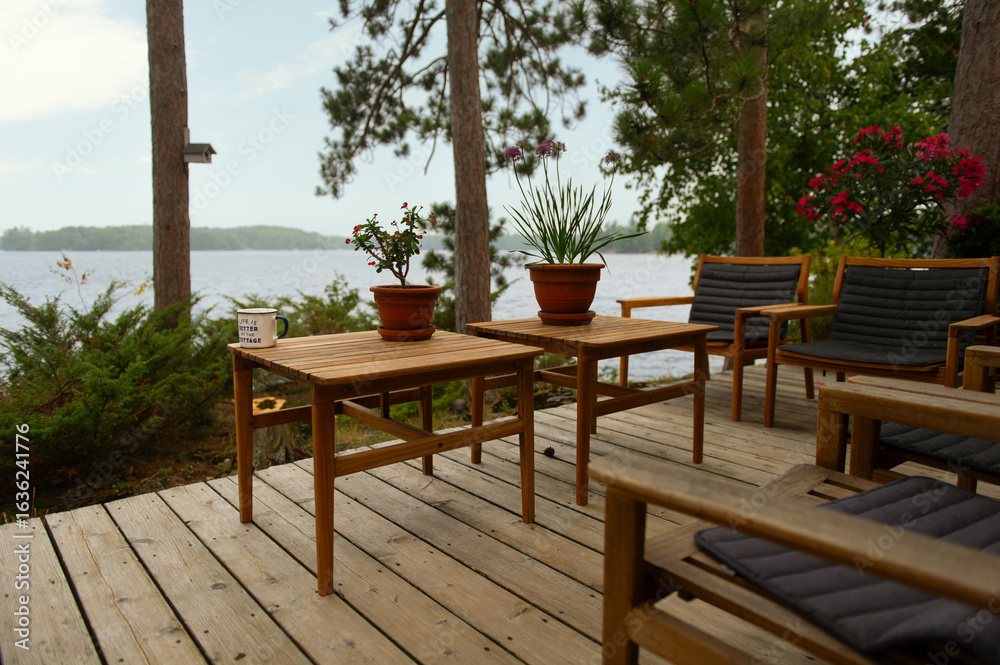 Obraz premium An outdoor wooden table and chairs sit on a deck surrounded by pines, overlooking a shimmering lake – an inviting setting for summer meals and long conversations in cottage country.