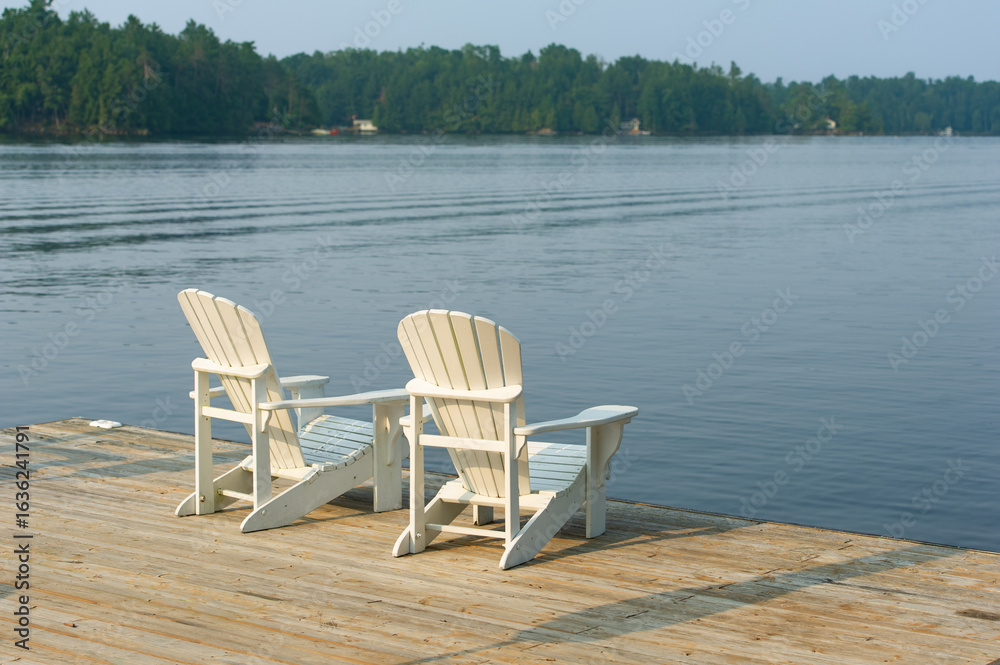 Fototapeta premium Minimalist white Adirondack chairs sit on a dock overlooking a mirror-like lake, the forest horizon glowing in the soft light of early morning – a tranquil retreat in Ontario's Kawarthas.