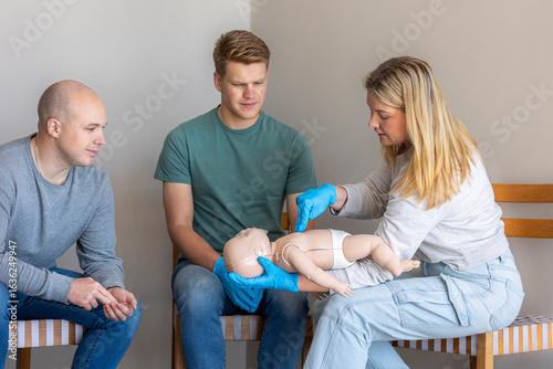 Female first aid instructor demonstrating the correct method for administering chest compressions on an infant Basic Life Support (BLS) CPR manikin, with students observing.