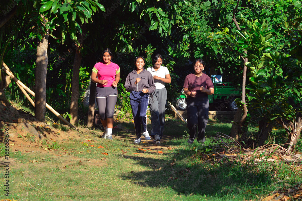 Obraz premium A group of four teen girls jog together across a lush tropical park in the morning, surrounded by green trees and sunlight filtering through the leaves.