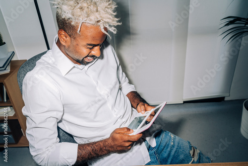 Direct flash shot of cheerful Middle Eastern man in white shirt with blond dreadlocks smiling and playing videogame on handheld gaming console while sitting at desk during break in work in office.