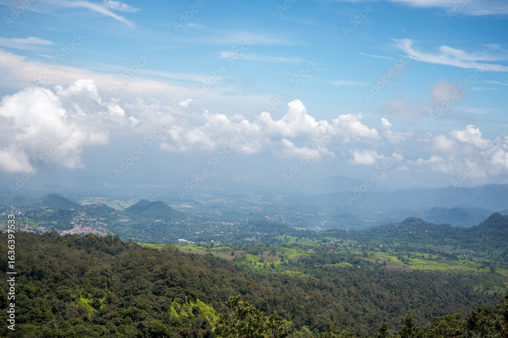 Naklejka premium Panoramic view of a lush green valley and distant mountains under a blue sky with scattered clouds.