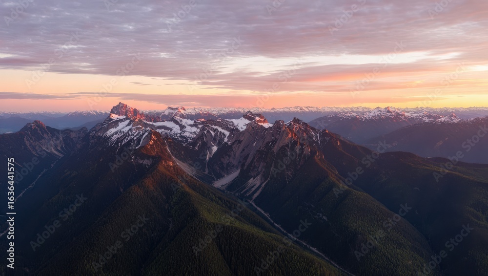 Naklejka premium Aerial view of snow capped mountains during sunset with a colorful sky and green forested valleys