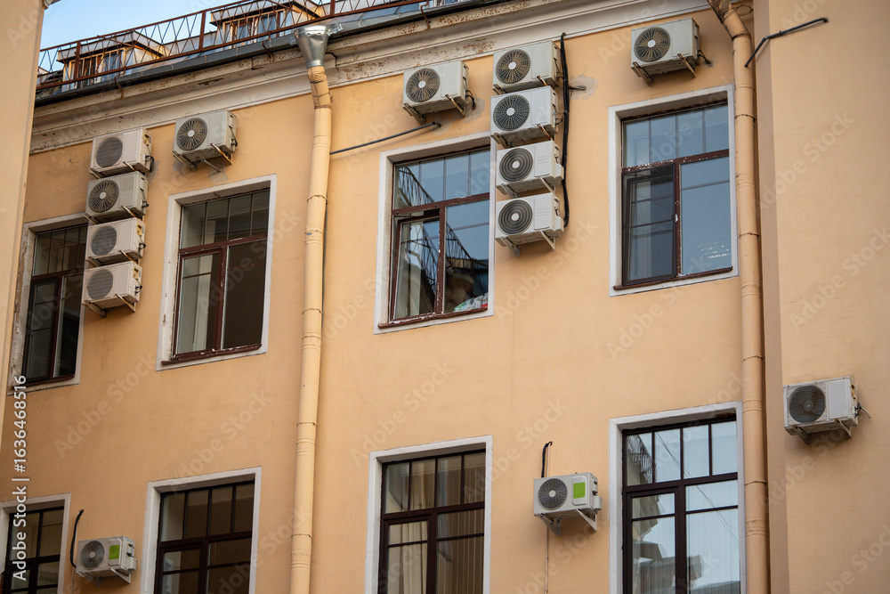 Obraz premium Facade of building with many air conditioners on windows in southern hot country. Yellow house exterior on city street. Escape from heat, creating favorable microclimate in urban apartments concept.