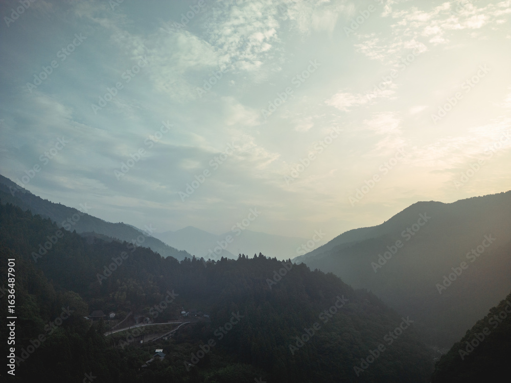 Fototapeta premium A serene landscape featuring mountains and a valley shrouded in mist. The sky is pale with wispy clouds, hinting at sunrise. A winding road cuts through the valley.