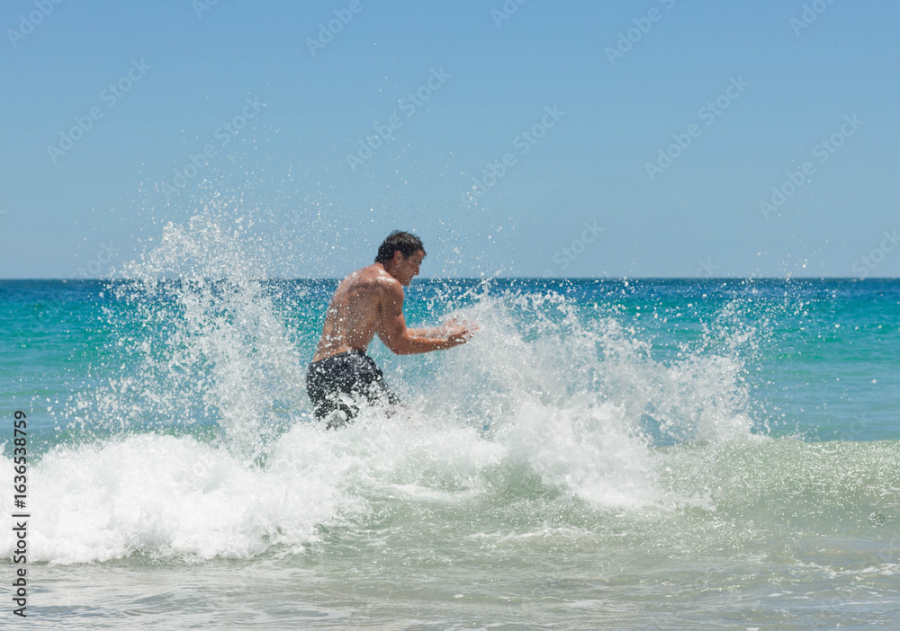 Obraz premium Man in dark swim shorts is wading in crashing turquoise waves at beach, sending water spray
