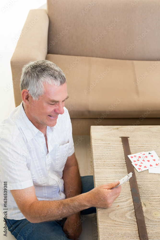 Fototapeta premium Senior man sitting holding cards in living room by coffee table with beige couch, copy space