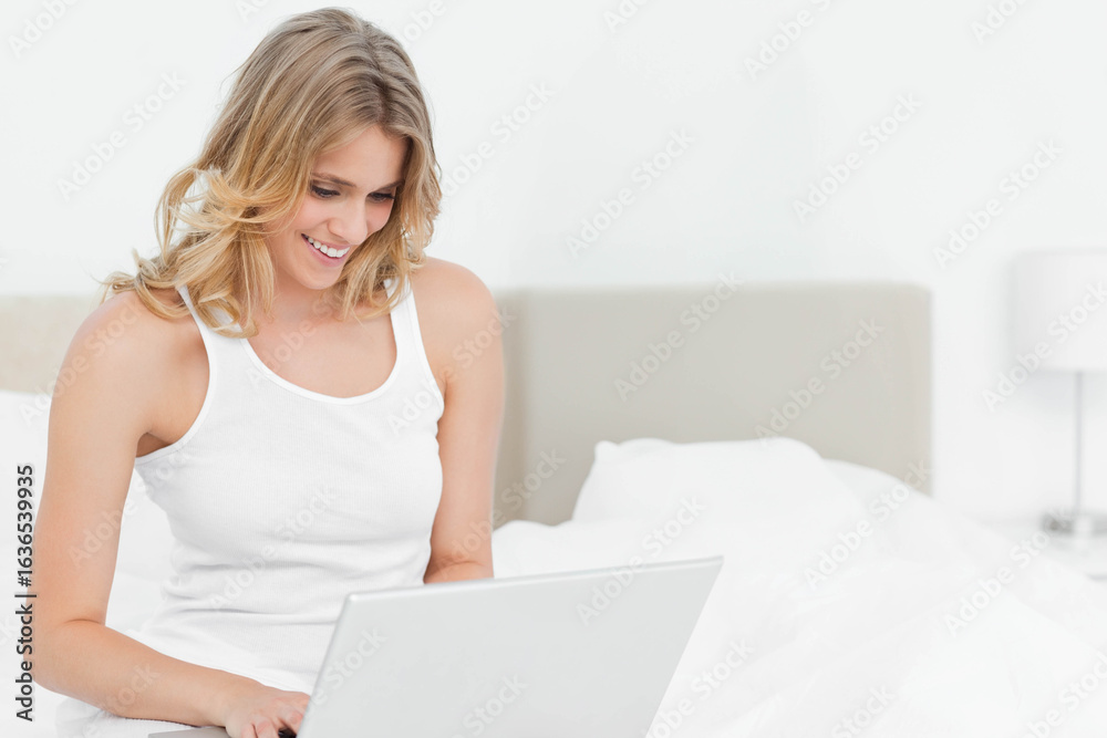 Woman sitting on bed in bedroom using laptop wearing tank top by lamp, copy space Woman sitting on bed in bedroom using laptop wearing tank top by lamp, copy space