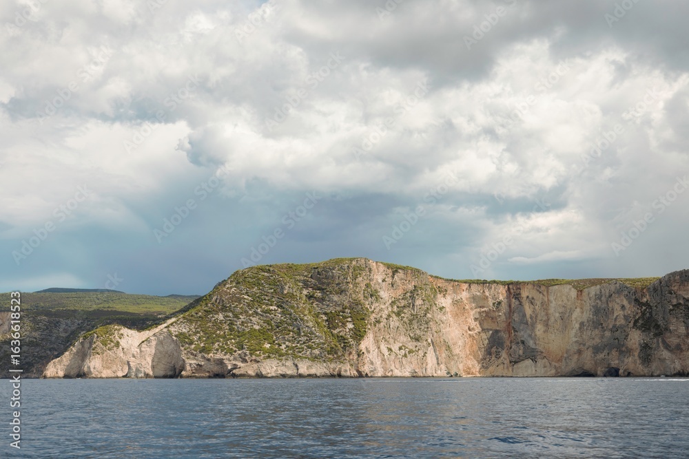 Fototapeta premium A coastal landscape in Greece shows a dramatic cliff face rising from the Ionian Sea under a sky filled with gathering storm clouds on a bright, overcast day