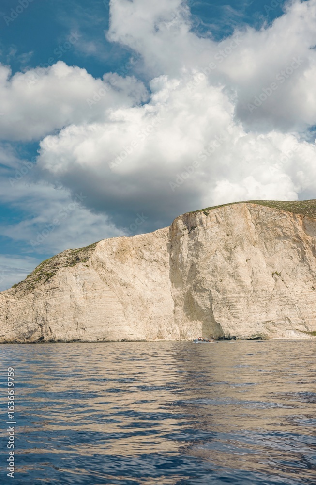 Naklejka premium A towering cliff face descends to the ocean, reflecting light in ripples. A vast cloudscape above dominates, offering a sense of scale in Zakynthos, Greece