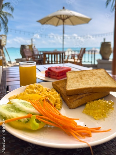 A low-angle shot captures a delicious breakfast served on a wooden table at a tropical beachside cafe. In the foreground, a white plate holds scrambled eggs, cucumber slices, shredded carrots, and two