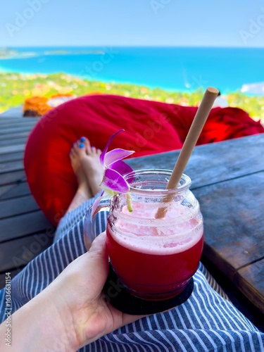 A first-person, low-angle shot shows a woman's hand holding a vibrant pink tropical drink in a glass jar with a straw and an orchid garnish. The woman's feet with blue painted nails are visible