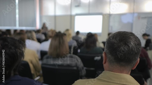 Man is sitting in a classroom with many other people. The man is looking at a projector screen