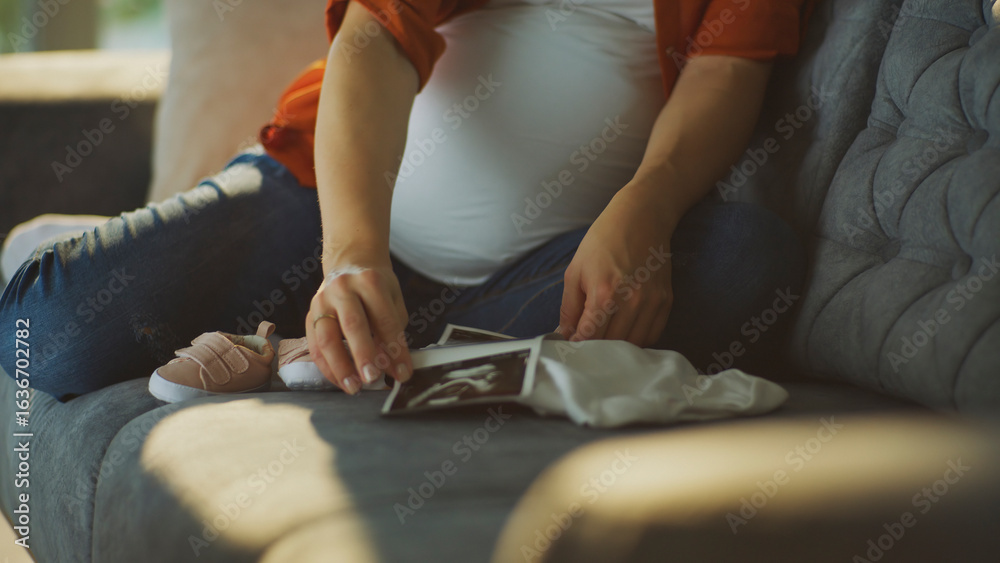 Fototapeta premium Pregnant woman holding ultrasound scan with baby clothes and shoes